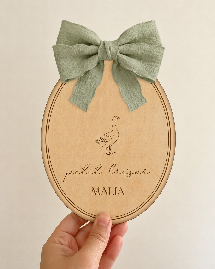 Plaque de naissance ovale gravée tenue à la main, nœud vert sauge passé dans la fente
